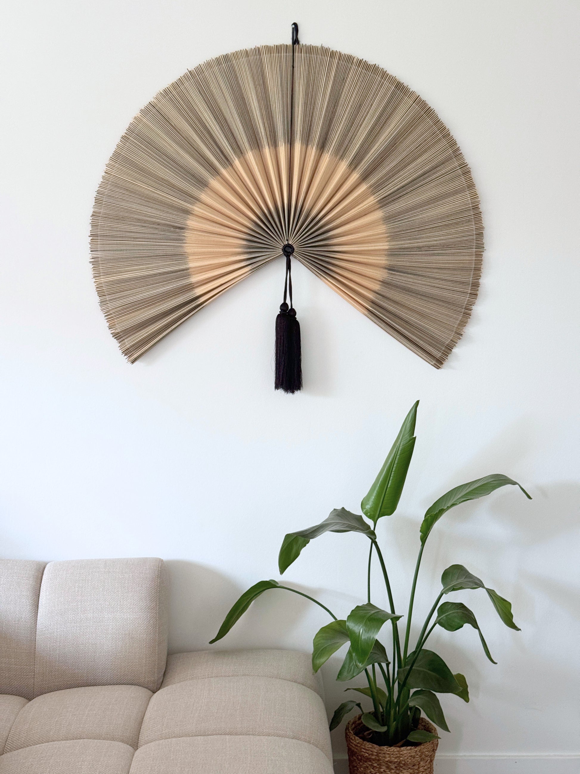 Håndlavet bambusvifte i naturlige beige nuancer med sort kvast, ophængt på en hvid væg over en lys sofa og grøn plante