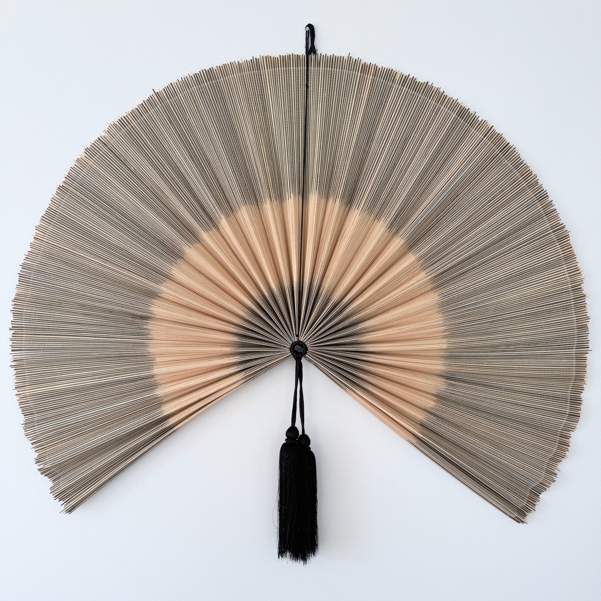 Håndlavet bambusvifte i naturlige beige nuancer med sort kvast, ophængt på en hvid væg