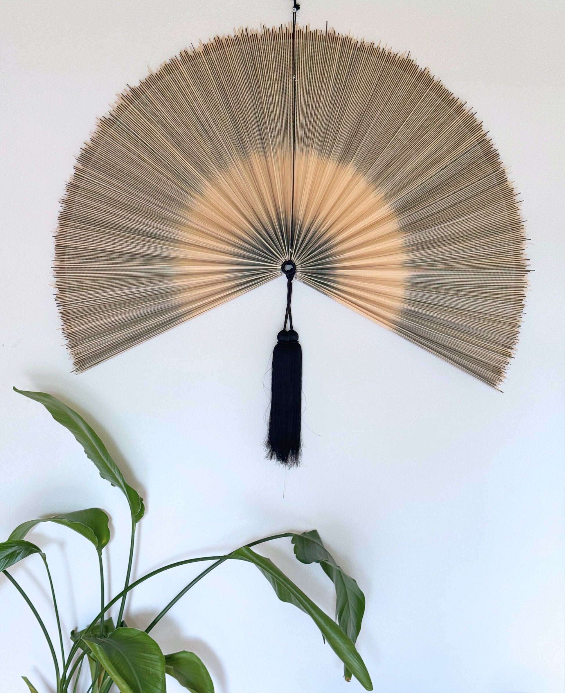 Håndlavet bambusvifte i naturlige beige nuancer med sort kvast, ophængt på en hvid væg