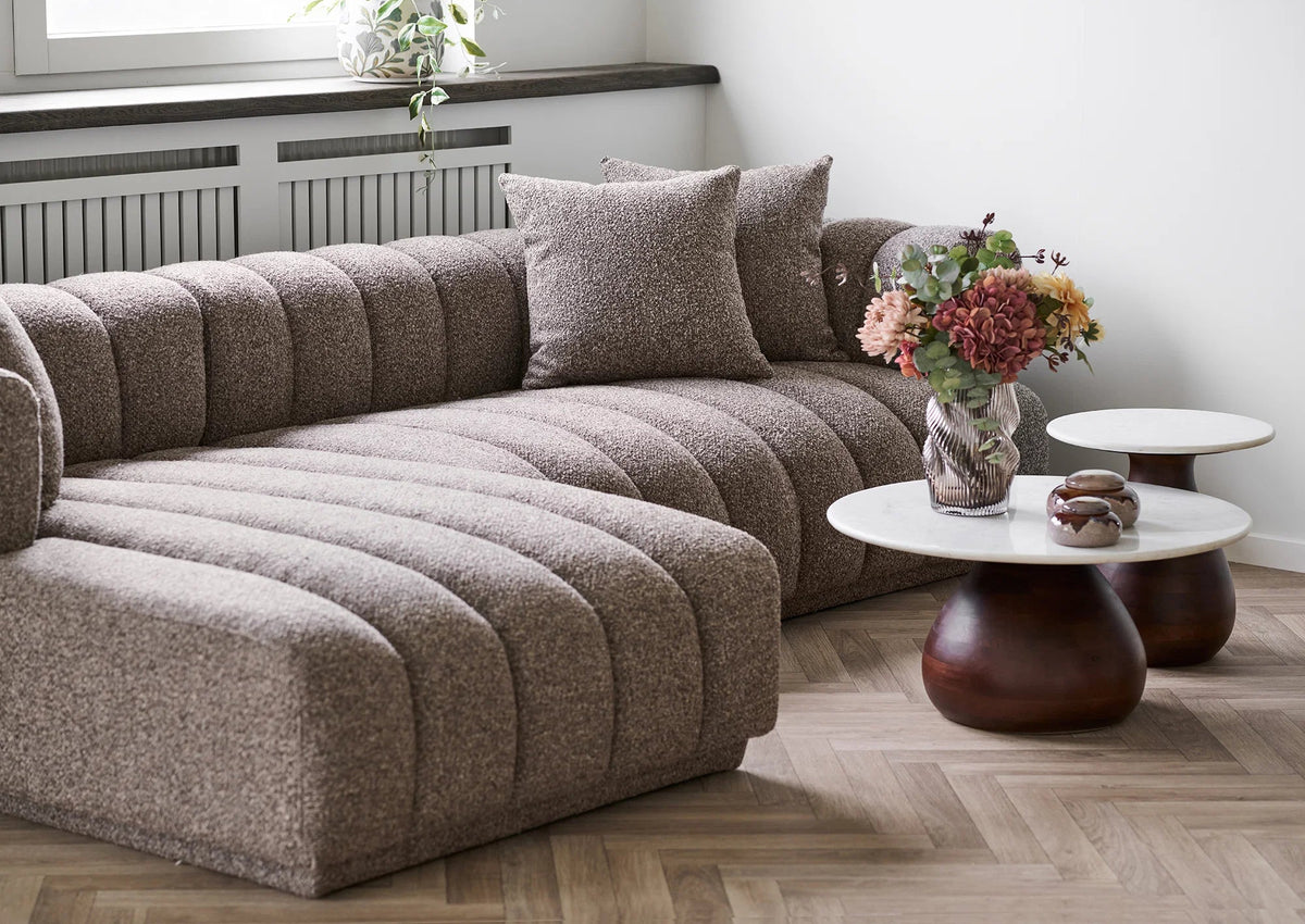 Aurora chaiselongsofa i brun set skråt forfra i en stue med blomster og runde sofaborde.