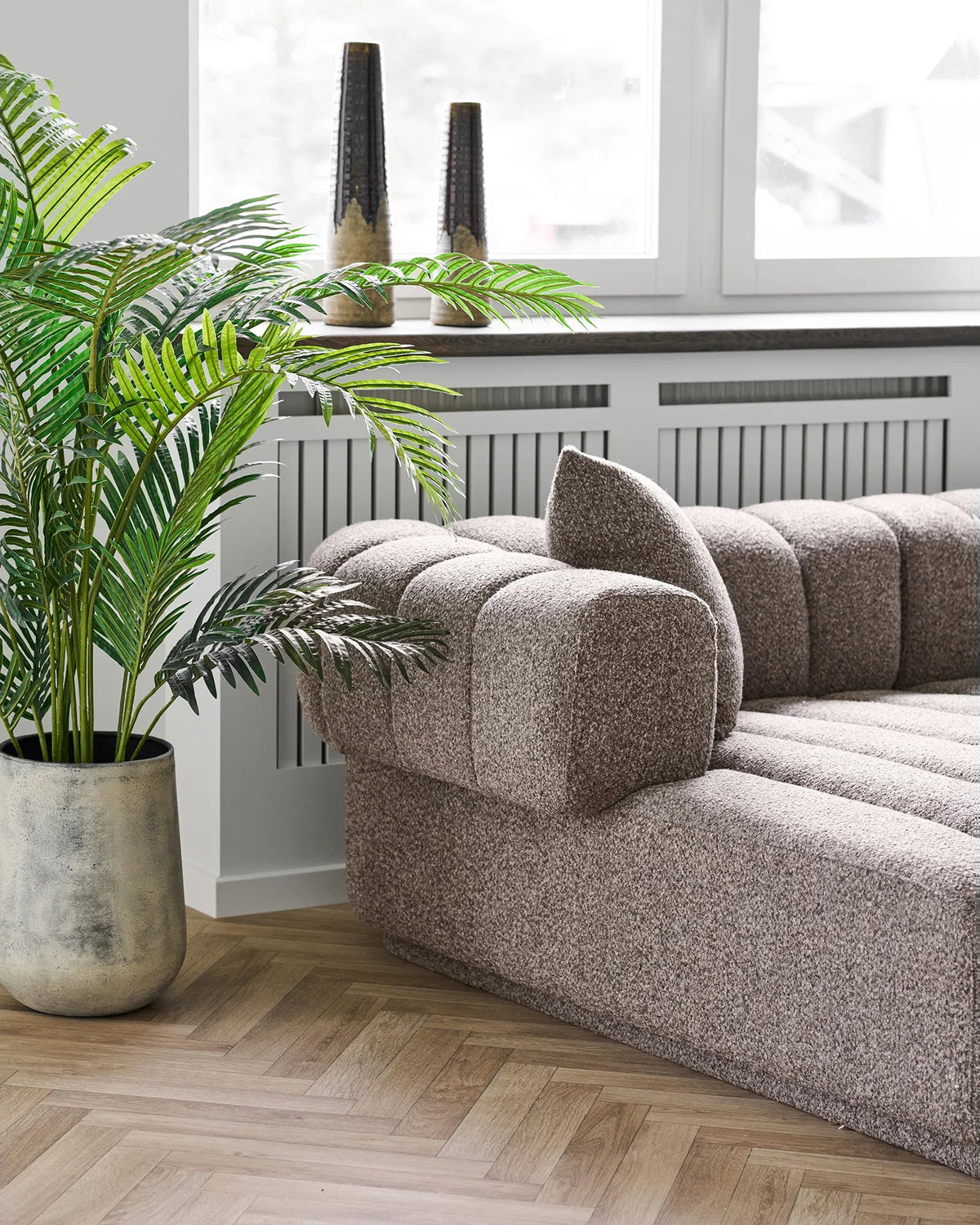 Aurora sofa i brun, chaiselong og afrundet armlæn set fra siden ved en grøn plante og et stort vindue.