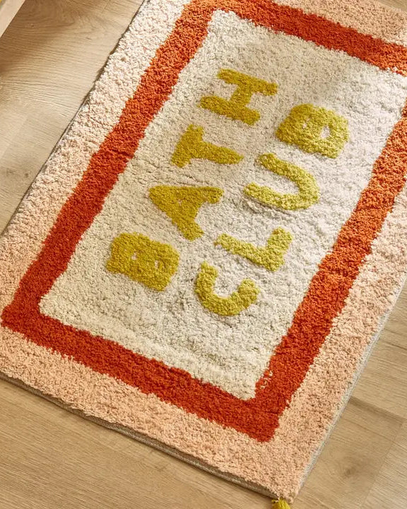 Blød bademåtte med teksten "Bath Club" i gul, creme og orange nuancer
