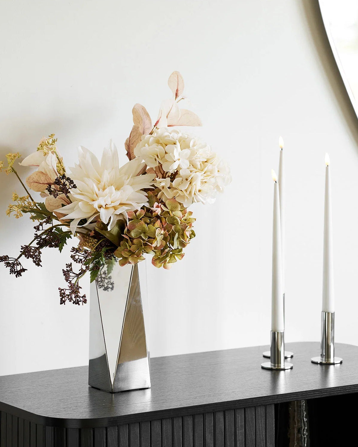 Blank vase i stål med blomsterbuket på mørkt skab, stylet med slanke lysestager og organisk vægspejl.
