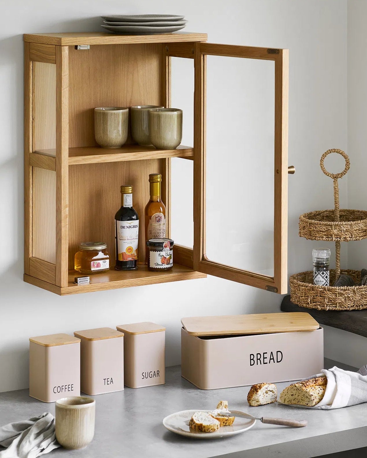 Eik væghængt vitrineskab i egetræ stylet i et lyst køkken med åben låge. Skabet er fyldt med keramikkopper, olier og marmelade.