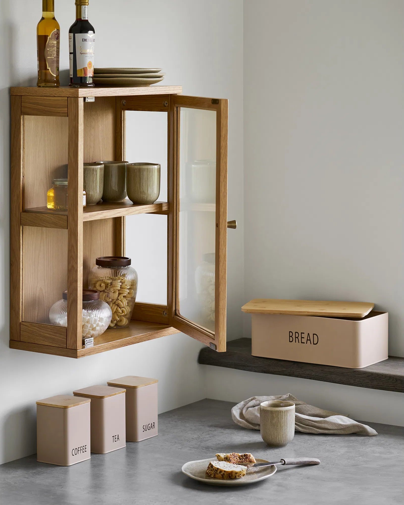 Eik væghængt vitrineskab hængende over et køkkenbord med en brødkasse, kaffedåser og friskbagt brød i en hyggelig nordisk indretning.