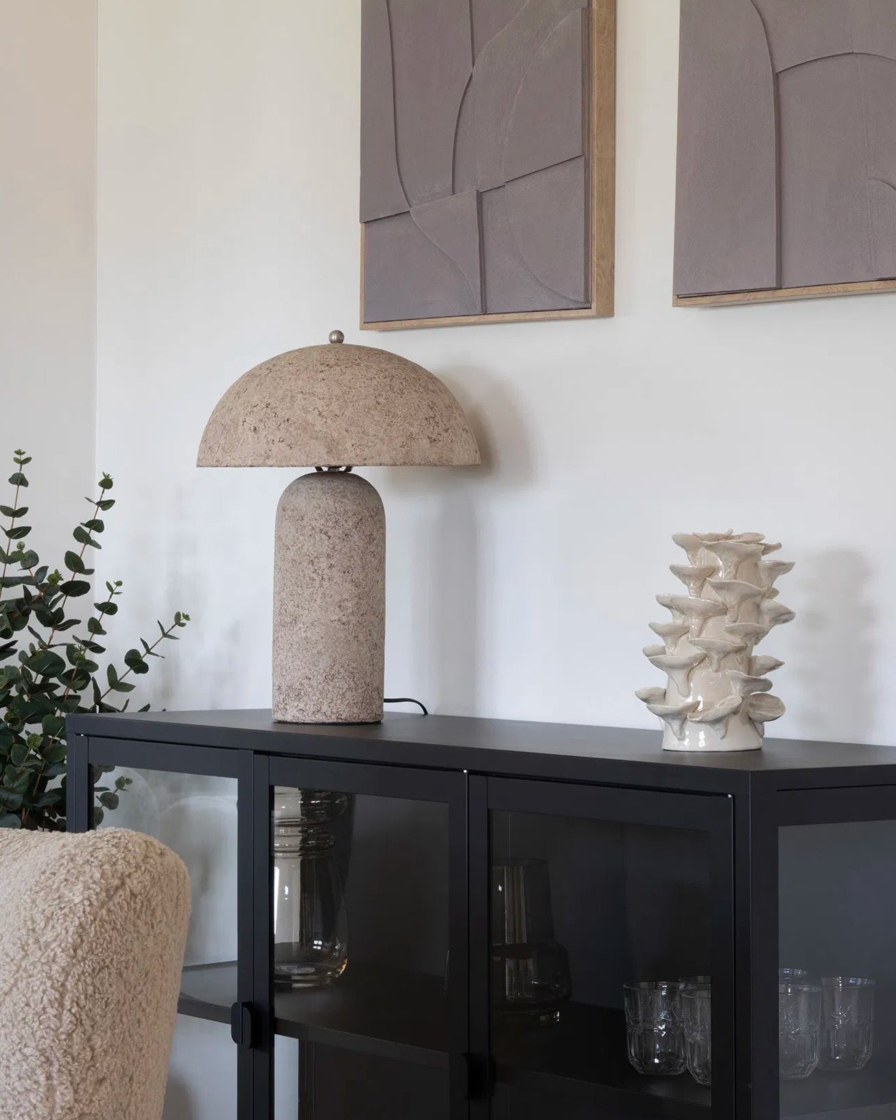 Molo bordlampe stylet på en sort metalskænk i en lys stue, sammen med en hvid keramikvase og grønne grene.