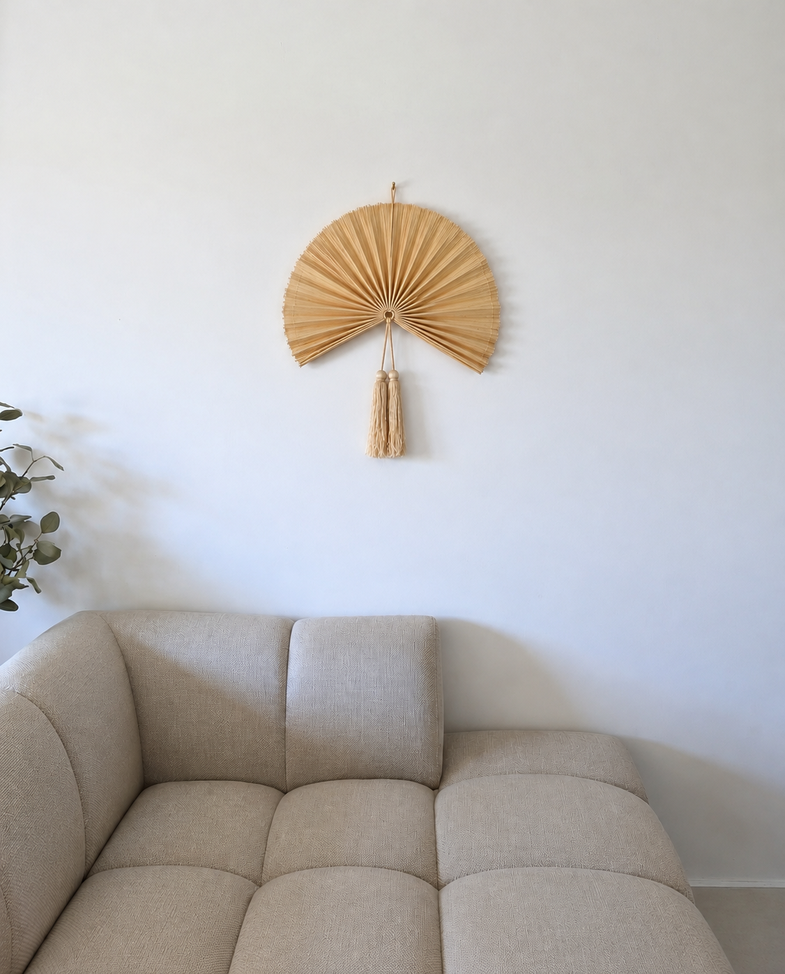 Lille gylden bambusvifte i naturfarvet bambus med dekorative kvaster, hængende på hvid væg over beige sofa i minimalistisk stue.