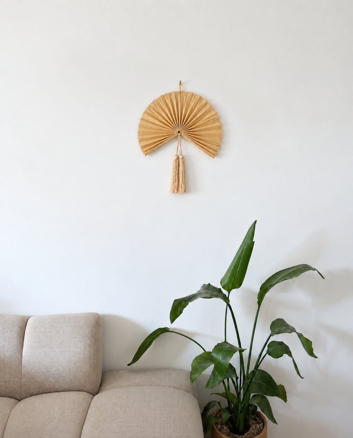 Lille gylden bambusvifte med lyse kvaster som vægdekoration over beige sofa ved siden af grøn plante i skandinavisk stue.