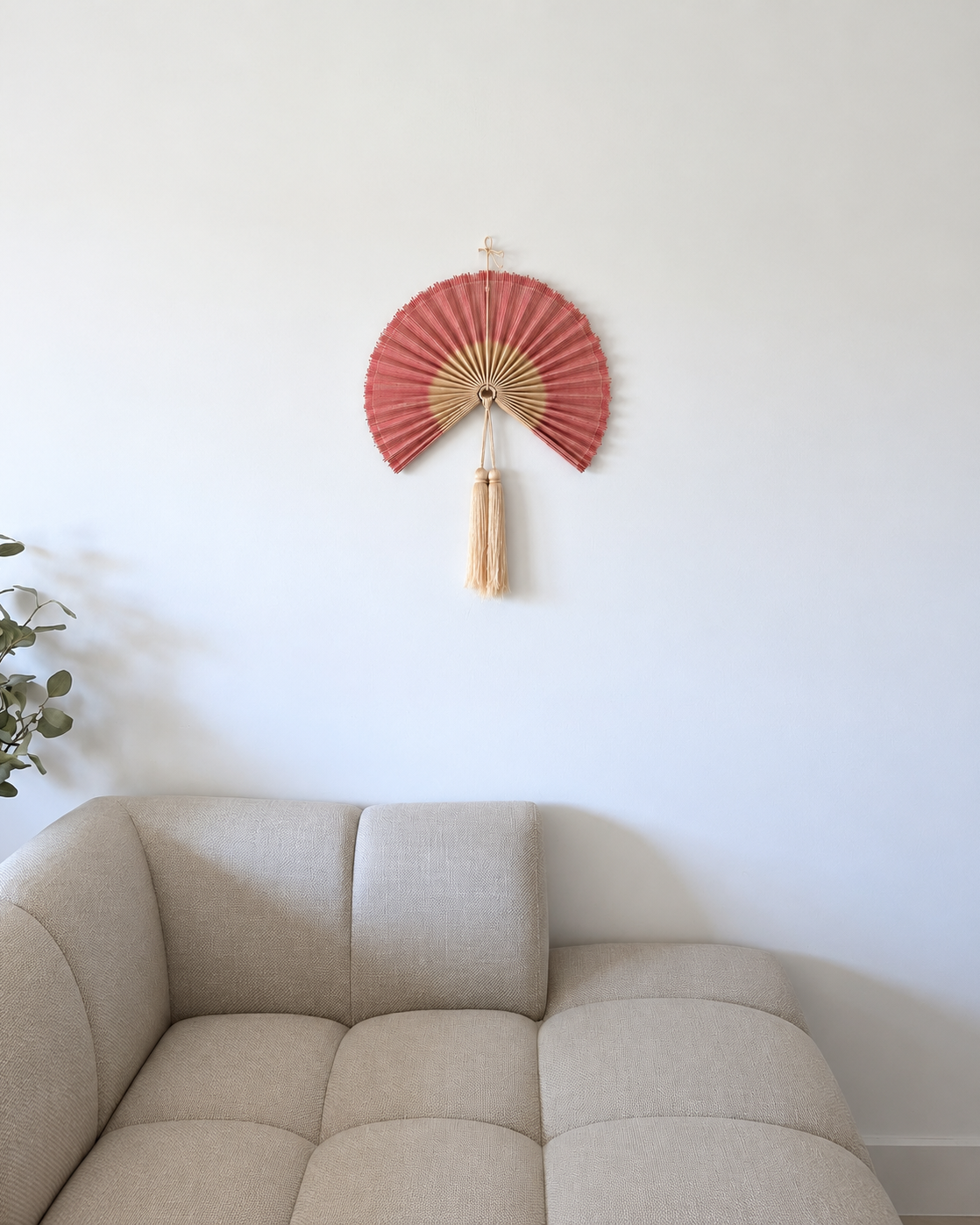 Lille rosa bambusvifte med naturfarvede detaljer og dekorative kvaster, hængende på hvid væg over beige sofa i nordisk stue.