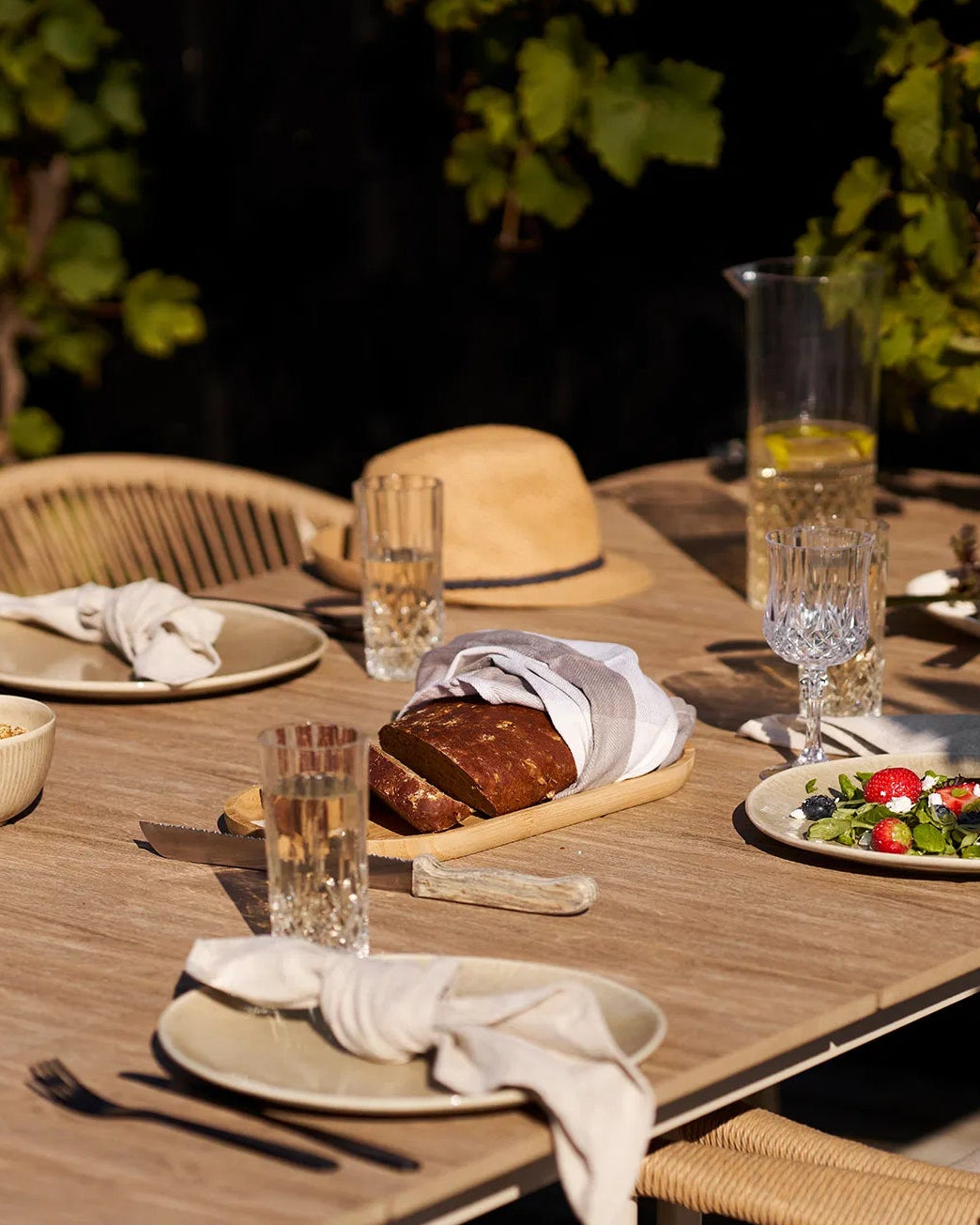 Nærbillede af dækket havebord med Kyst middagstallerkener, hørservietter og krystalglas i sommersolskin.