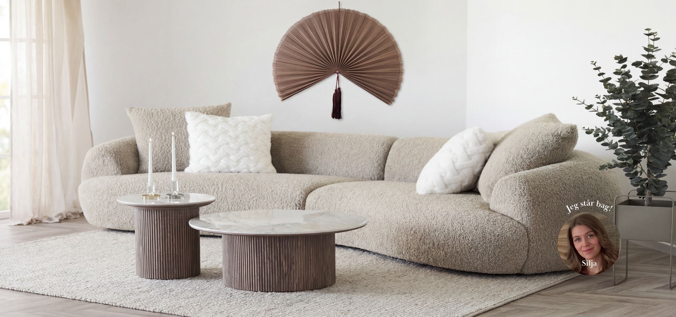 Stue i lyse, nordiske farver indrettet med en stor, beige bouclé sofa, runde sofaborde og en brun Saga bambusvifte som vægdekoration fra Nilsson Living.