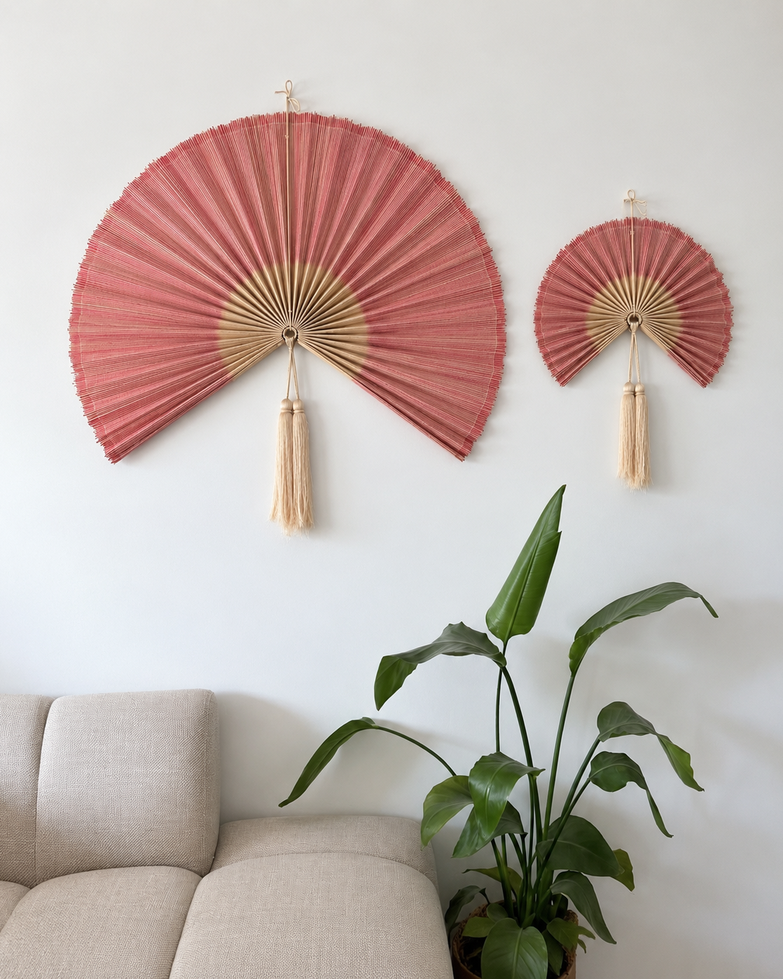 Stor og lille rosa bambusvifte med naturfarvede detaljer og lange kvaster, hængende side om side over beige sofa i moderne stue.