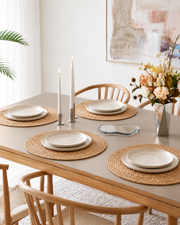 Fire ovale flettede dækkeservietter i naturfarve på spisebord med beige tallerkener, levende lys og blomster i skandinavisk indretning.