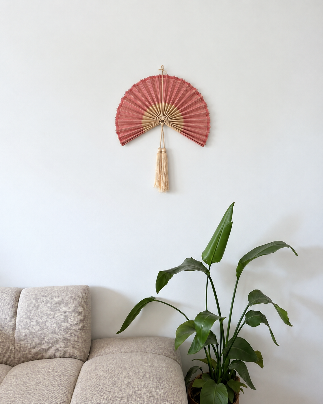 Lille rosa bambusvifte som dekorativ vægpynt over beige sofa ved siden af grøn plante i lys, skandinavisk stue.